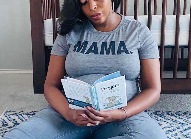 African American pregnant woman with a gray shirt that says MAMA sitting on the floor reading a book titled Prayers for my Baby Boy.